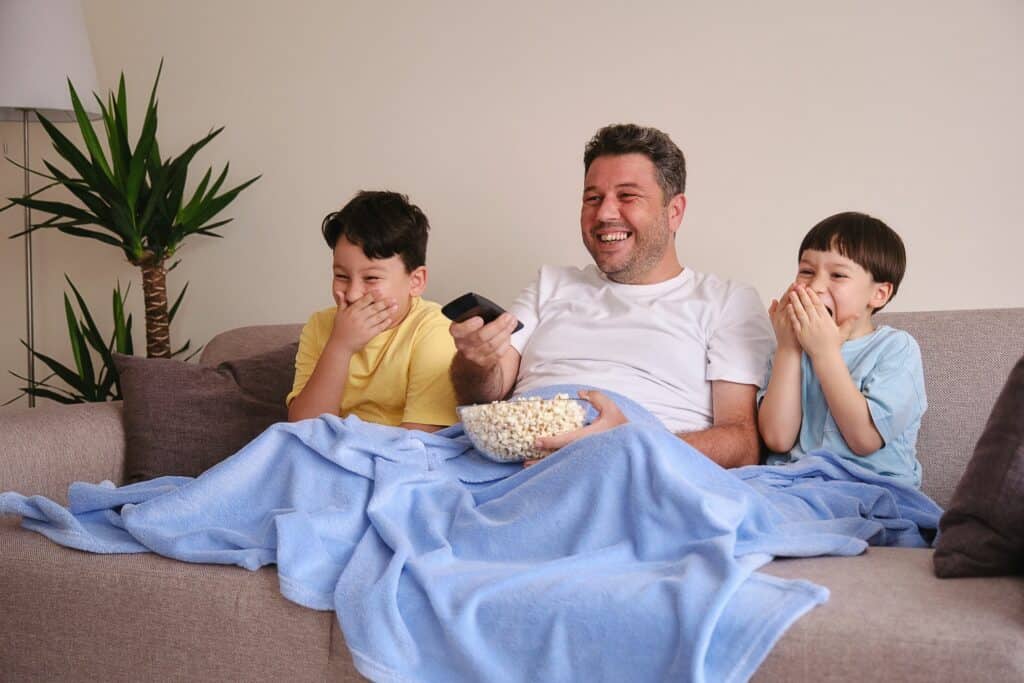 pai e filhos na sala assistindo tv com um balde de pipoca representando brincadeiras para dias de chuva