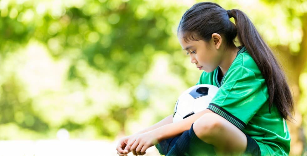 menina sentada em quadra de futebol segurando bola pensando em como lidar com a frustração