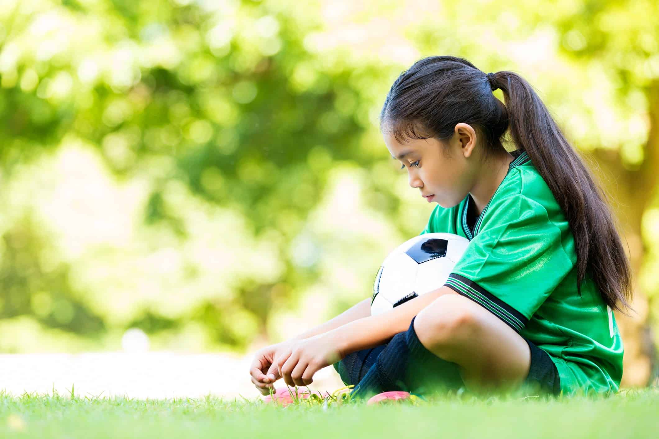 menina sentada em quadra de futebol segurando bola pensando em como lidar com a frustração