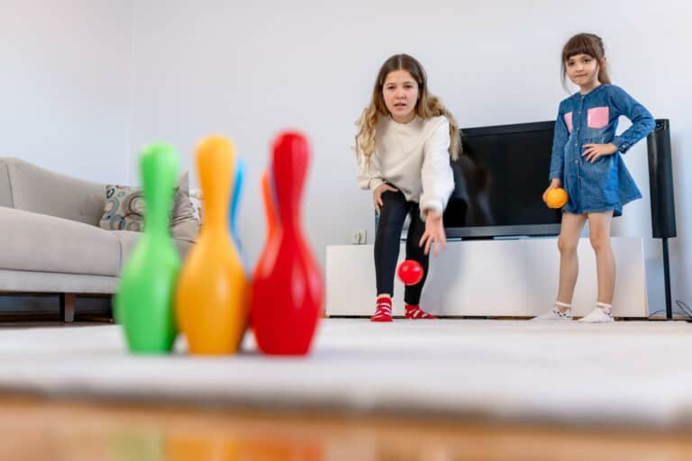 Imagem de duas crianças jogando boliche na sala de uma casa. O brinquedo tem pinos coloridos.