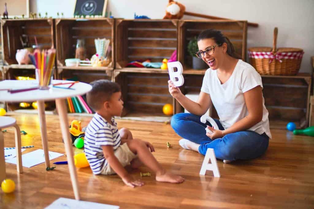 Imagem de uma mulher ensinando o alfabeto para uma criança em uma sala de estar.