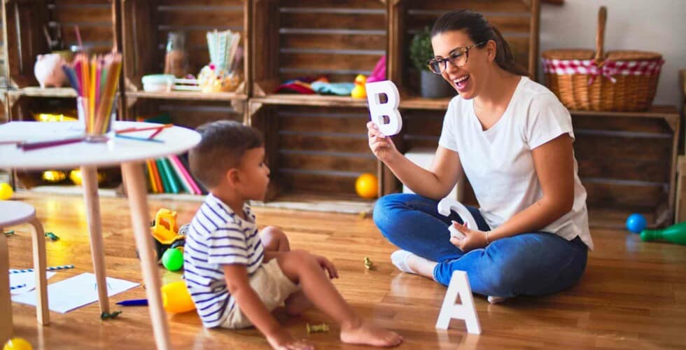 Imagem de uma mulher ensinando o alfabeto para uma criança em uma sala de estar.