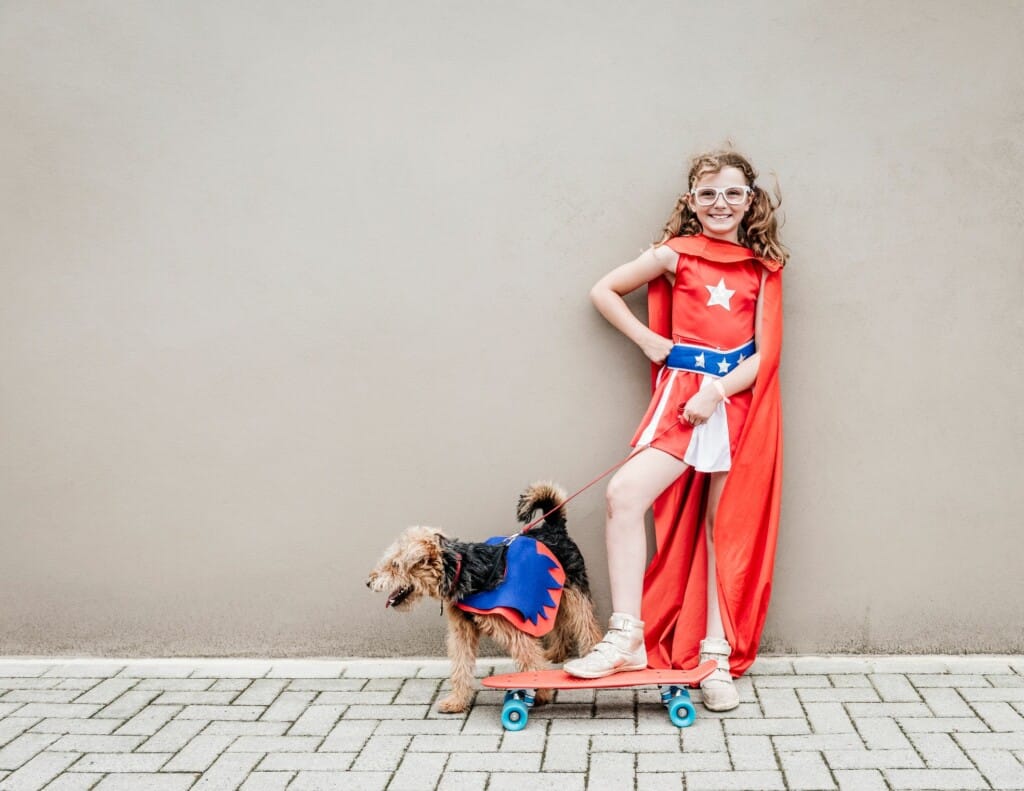 Imagem de uma criança vestida de super heroína ao lado de um cachorro.