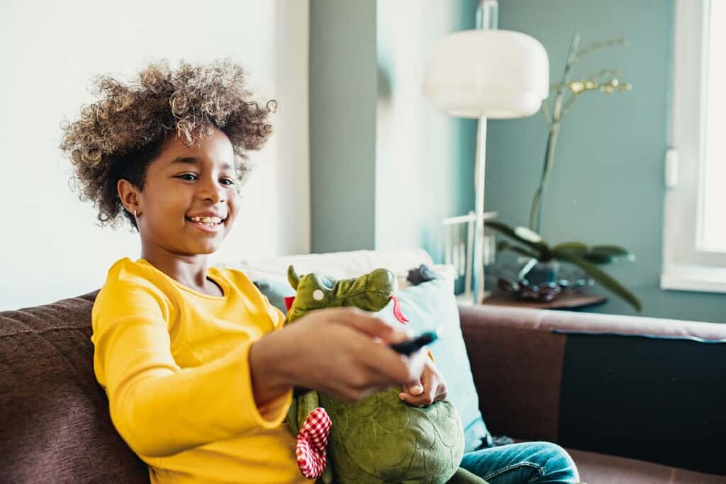 Imagem de uma criança segurando um controle remoto sentada num sofá.