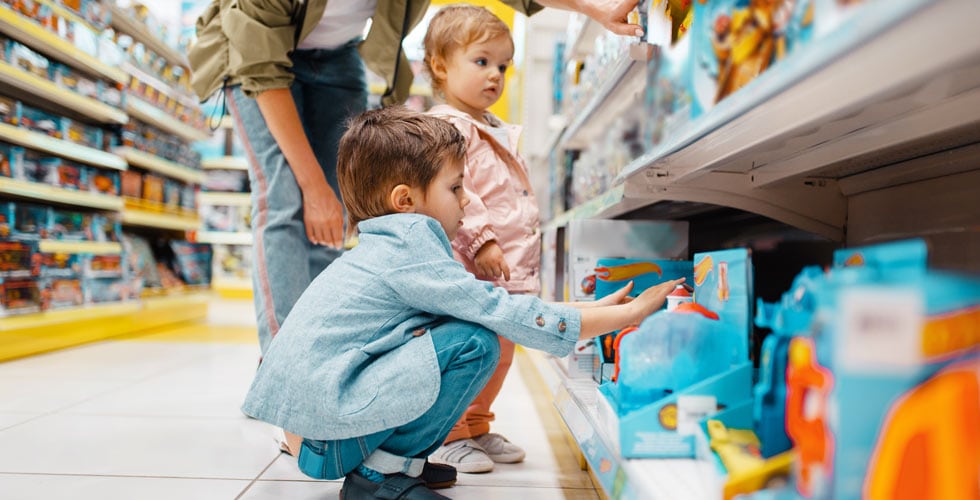mãe ao lado do filho e filha em frente a prateleira com brinquedos representando o consumo infantil