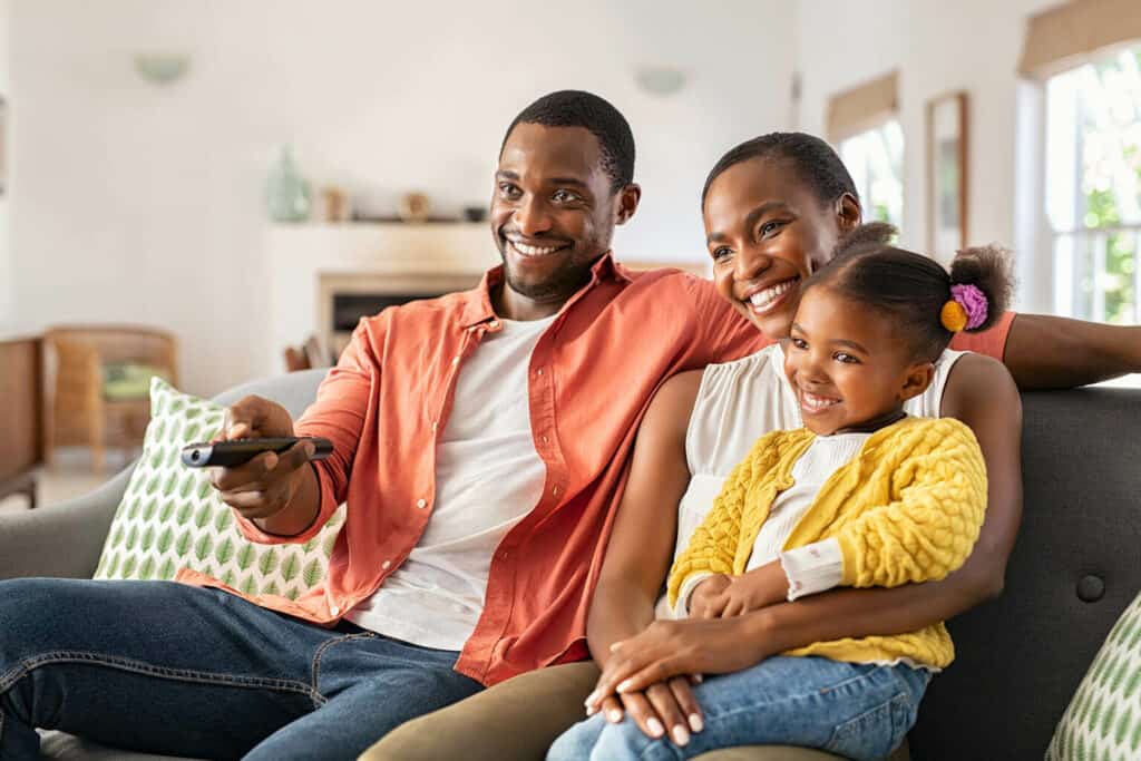 Imagem de dois adultos e uma criança sorrindo ao assistir a televisão