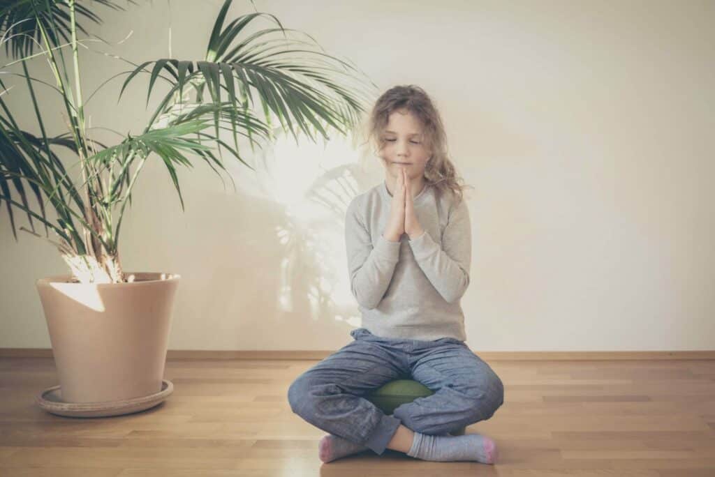 menina sentada em almofada com as mãos unidas representando a meditação para crianças 