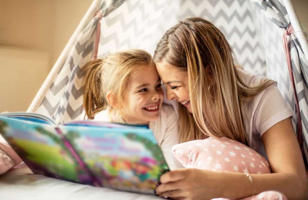 mãe e filha sorrindo uma para outra enquanto leem juntas em cima da cama representando o fortalecimento de vínculos entre pais e filhos