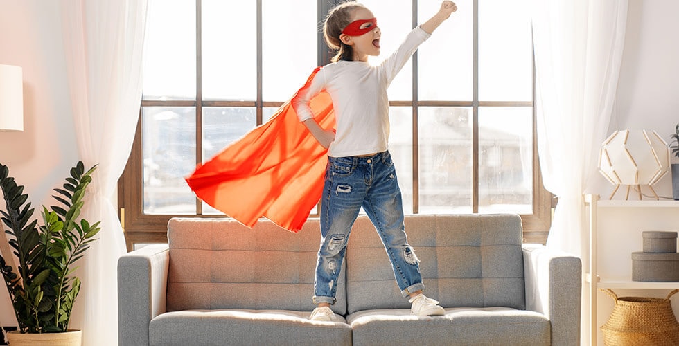Imagem de uma criança vestida de superherói e brincando em cima de um sofá