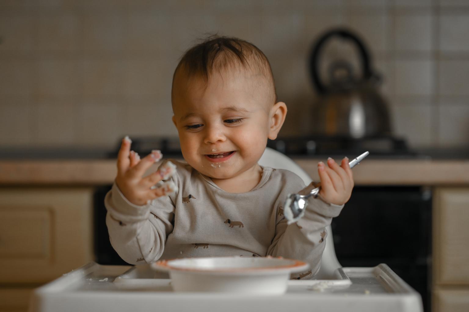 bebê sorridente sentado a mesa em frente a prato segurando colher representando a introdução alimentar