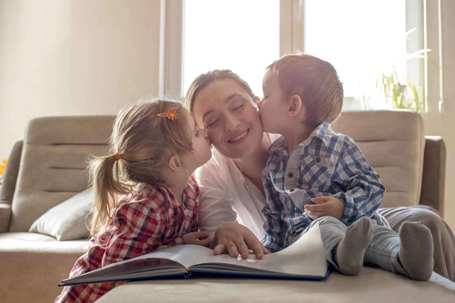 crianças beijando o rosto de mulher adulta em frente a mesa com livro representando o dia do filho